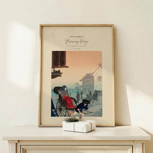 May include: Framed Japanese woodblock print showing a woman in red kimono sitting in a rickshaw with a man kneeling beside it, set against peachy sunset sky with traditional buildings. Natural wood frame with cream matting displays the vintage-style artwork on light wooden furniture next to small white wrapped gift with twine and greenery sprig.
