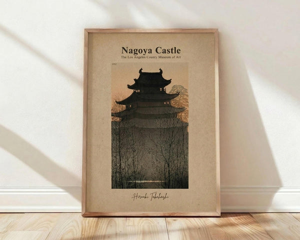 May include: Framed vintage-style poster showing a dark silhouette of a multi-tiered Japanese castle against a dusky orange-pink sky, surrounded by bare winter trees. The artwork has an aged, sepia-toned background with text reading "Nagoya Castle" and "The Los Angeles County Museum of Art" at the top, "1952" in the middle, and a signature at the bottom. The wooden frame sits on light wood flooring against a white wall with natural lighting creating soft shadows.