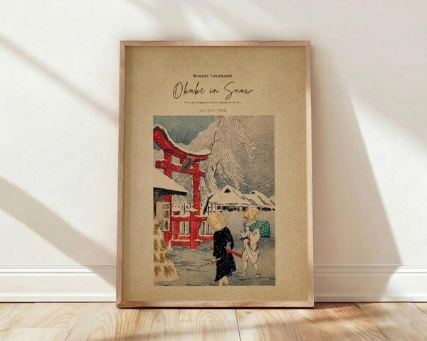 May include: Framed vintage Japanese woodblock print showing figures with umbrellas walking past a red torii gate in falling snow, with Mount Fuji in the background. The artwork features muted colors on cream matting within a light wood frame, positioned against white walls on light wooden flooring with soft natural lighting.
