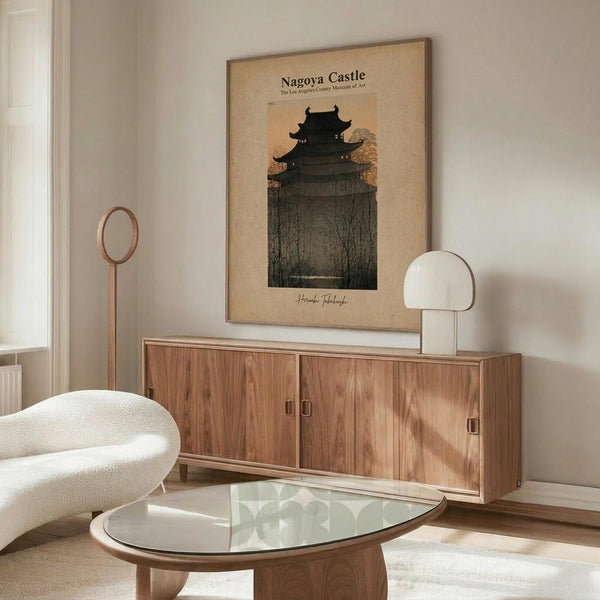 May include: A framed vintage-style poster featuring a dark silhouetted Japanese pagoda castle against a warm beige background with bare tree branches. The artwork hangs above a mid-century modern walnut credenza with sliding doors in a minimalist living room setting. A white curved lounge chair sits beside an oval glass coffee table with wooden base, while a sleek table lamp and floor lamp provide ambient lighting on neutral walls.