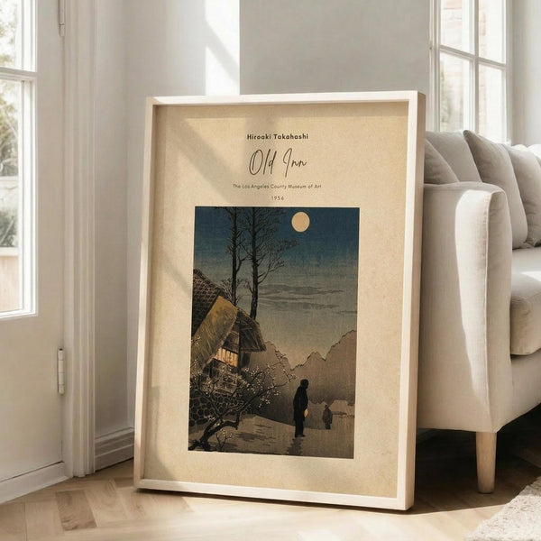 May include: Framed vintage Japanese woodblock print poster leaning against white wall next to beige sofa. Shows moonlit winter scene with traditional thatched roof house, bare tree, two silhouetted figures walking on snowy path, and mountains in background. Cream colored mat with museum exhibition text above artwork in light wood frame.