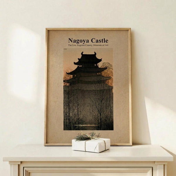 May include: Framed vintage-style poster featuring Japanese castle silhouette against twilight sky with bare winter trees in foreground, displayed on light wood furniture beside small white wrapped gift with greenery sprig. Warm beige tones throughout scene with natural lighting creating soft shadows on white wall.