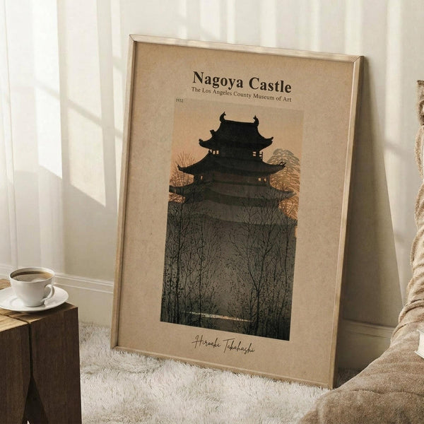 May include: Framed vintage-style poster featuring a dark silhouette of a traditional Japanese castle with multiple tiers and curved roofs, surrounded by bare winter trees. The artwork has muted earth tones with a beige background. The frame leans against a white paneled wall next to a wooden side table with a white coffee cup, and a cream knitted throw blanket is draped nearby on what appears to be a white textured rug.