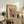 May include: Framed vintage Japanese woodblock print poster featuring a snowy mountain scene with red torii gate, traditional buildings, and figures in winter clothing. Displayed on rustic wooden sideboard alongside fern plant, ceramic vase, books, and warm neutral home decor. Natural lighting creates cozy living room atmosphere with beige walls and brown leather furniture visible.