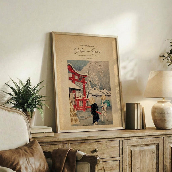May include: Framed vintage Japanese woodblock print poster featuring a snowy mountain scene with red torii gate, traditional buildings, and figures in winter clothing. Displayed on rustic wooden sideboard alongside fern plant, ceramic vase, books, and warm neutral home decor. Natural lighting creates cozy living room atmosphere with beige walls and brown leather furniture visible.