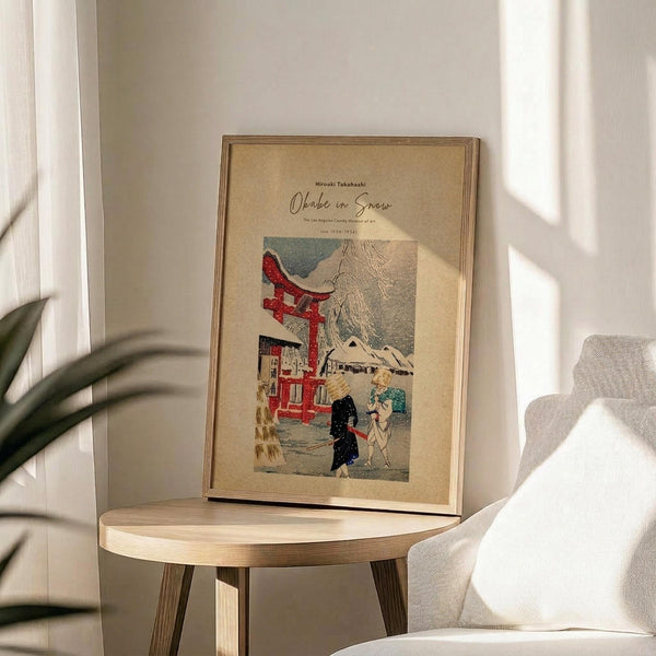 May include: Framed vintage Japanese woodblock print poster featuring a red torii gate, snow-covered buildings, and figures in winter scene. Warm beige matting in thin wooden frame, displayed on round wooden side table next to cream upholstered chair. Soft natural lighting creates shadows across neutral-toned room with plant visible in corner.