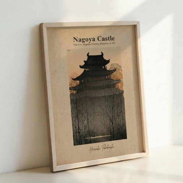 May include: Framed vintage-style Japanese woodblock print poster showing Nagoya Castle's dark silhouette against golden sunset sky, surrounded by bare winter trees. Features cream-colored mat with museum attribution text and artist signature. Light wood frame leans against white wall, casting soft shadows. Warm, muted earth tones throughout the atmospheric twilight scene.