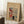 May include: Framed vintage Japanese woodblock print poster leaning against white wall on white textured rug. Print shows snowy winter scene with red torii gate, figures walking with umbrellas, and mountain in background on tan paper. Natural wood frame, displayed next to wooden side table with white coffee cup and saucer, cozy blanket visible at edge.