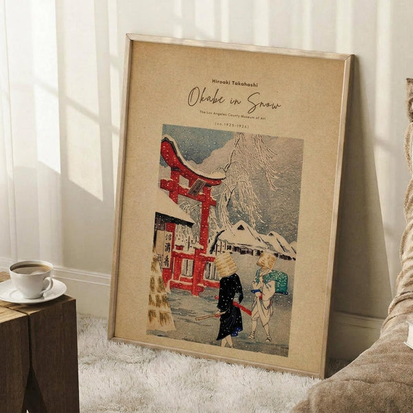 May include: Framed vintage Japanese woodblock print poster leaning against white wall on white textured rug. Print shows snowy winter scene with red torii gate, figures walking with umbrellas, and mountain in background on tan paper. Natural wood frame, displayed next to wooden side table with white coffee cup and saucer, cozy blanket visible at edge.