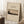 May include: Vintage-style poster featuring Mount Fuji with snow-capped peak rising behind dark silhouetted trees in muted earth tones, displayed in light wood frame leaning against white wall near window, with coffee cup on wooden block and cozy textured blanket, creating serene minimalist room setting with natural lighting.