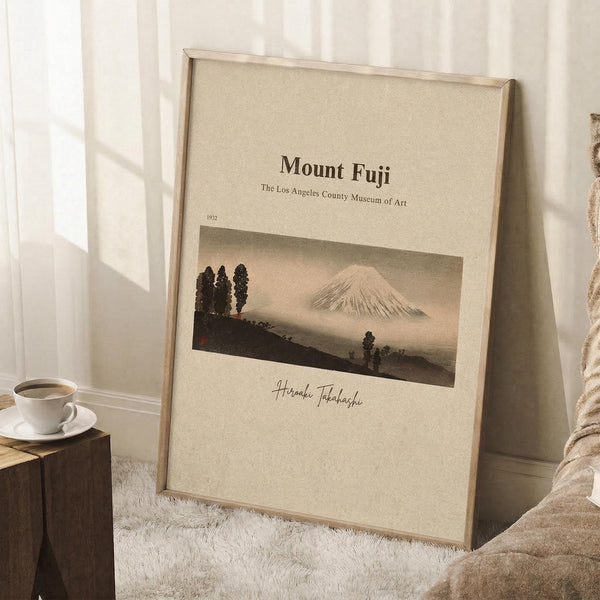 May include: Vintage-style poster featuring Mount Fuji with snow-capped peak rising behind dark silhouetted trees in muted earth tones, displayed in light wood frame leaning against white wall near window, with coffee cup on wooden block and cozy textured blanket, creating serene minimalist room setting with natural lighting.