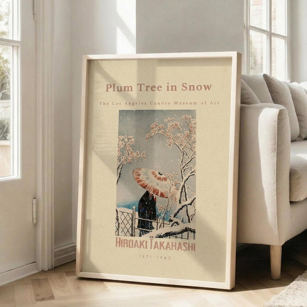 May include: Framed art print leaning against white wall next to beige sofa in bright living room. Print shows Japanese woodblock style winter scene with person holding red and white umbrella walking past snow-covered plum tree branches. Cream colored poster with museum exhibition styling, housed in light wood frame. Natural lighting creates soft shadows on hardwood floor.