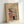 May include: Framed vintage Japanese woodblock print reproduction showing a snowy scene with red torii gate, traditional buildings, and two figures in winter clothing walking through falling snow. Print has cream-colored background with muted colors including red, blue, and black. Displayed in light wood frame leaning against white wall on white surface.