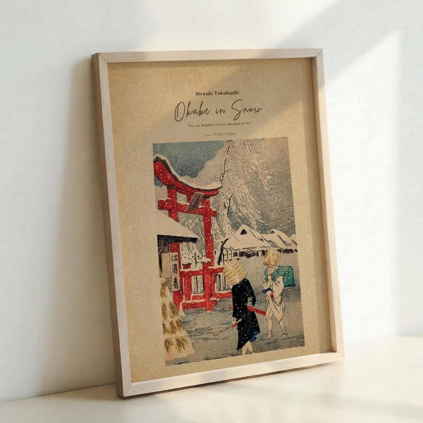 May include: Framed vintage Japanese woodblock print reproduction showing a snowy scene with red torii gate, traditional buildings, and two figures in winter clothing walking through falling snow. Print has cream-colored background with muted colors including red, blue, and black. Displayed in light wood frame leaning against white wall on white surface.