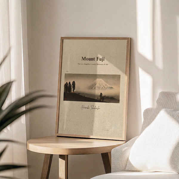 May include: Framed poster displaying Mount Fuji landscape artwork in muted earth tones, featuring the iconic mountain peak with trees in foreground and museum-style text layout. Light wooden frame sits on round wooden side table next to beige upholstered chair. Soft natural lighting creates gentle shadows across cream-colored wall, with green plant leaves visible in corner.