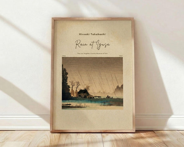 May include: Framed vintage Japanese woodblock print showing a rainy landscape scene with diagonal rain lines, traditional houses near water, trees, and mountains in muted earth tones of brown, green, and beige. The print is displayed in a light wooden frame leaning against a white wall on hardwood flooring with soft natural lighting creating shadows.