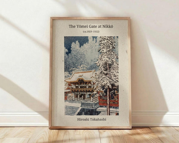 May include: Framed vintage Japanese woodblock print showing a snow-covered temple gate with traditional architecture, featuring red lacquered railings, ornate roof details, and snow-laden trees against a dark blue sky. The cream-colored mat displays the title and artist name in black text. Light wood frame sits on wooden flooring against white walls with soft natural lighting creating diagonal shadows.