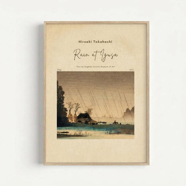 A framed vintage Japanese woodblock print reproduction showing a rainy landscape scene with diagonal rain lines across a muted sky. The artwork depicts a traditional house near a blue-green body of water, surrounded by dark silhouetted trees and rolling hills. The print has warm beige and brown tones with touches of turquoise water. Text appears above the image identifying the artist and artwork title. The piece is displayed in a light wooden frame against a white wall, creating soft shadows.