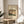 May include: Framed vintage Japanese woodblock print poster showing a rainy landscape scene with traditional buildings, trees, and water in muted earth tones and blues. The cream-colored print sits on a light wood round side table next to a beige upholstered chair, with plant leaves visible in soft natural lighting against white walls.