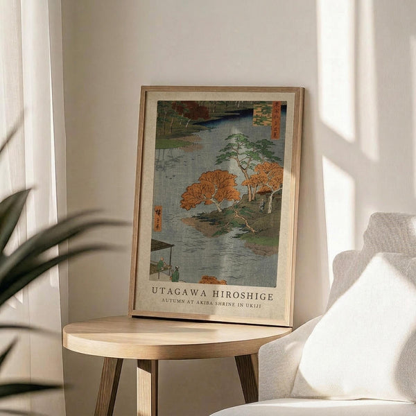 May include: Framed Japanese woodblock print featuring autumn scene with orange foliage trees beside flowing water, displayed on round wooden side table next to beige upholstered chair. Natural lighting creates soft shadows across white wall. Green plant leaves visible in corner. Print shows traditional ukiyo-e art style with muted blue-gray water and vibrant fall colors in light wood frame.