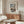 May include: Framed vintage Japanese woodblock print artwork hanging above a walnut sideboard in a minimalist living room. The print shows a snow-covered temple gate with traditional architecture in muted blues, whites, and earth tones. Modern white curved chair and glass-top coffee table visible in foreground, with contemporary table lamp and floor lamp completing the neutral-toned interior scene.