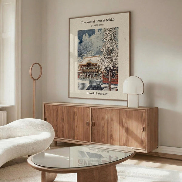 May include: Framed vintage Japanese woodblock print artwork hanging above a walnut sideboard in a minimalist living room. The print shows a snow-covered temple gate with traditional architecture in muted blues, whites, and earth tones. Modern white curved chair and glass-top coffee table visible in foreground, with contemporary table lamp and floor lamp completing the neutral-toned interior scene.