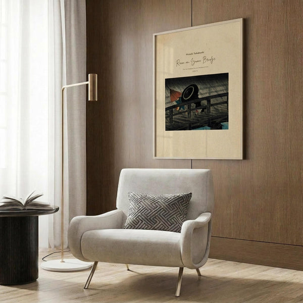 May include: Framed vintage Japanese woodblock print showing a bridge scene with rain, displayed on wood paneled wall above beige upholstered armchair with geometric patterned pillow. Modern reading corner with brass floor lamp and dark round side table holding open book. Warm neutral tones throughout staged interior setting.