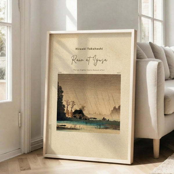 May include: Framed vintage Japanese woodblock print showing a rainy landscape with traditional house, blue water, bare trees, and diagonal rain lines against beige sky. Light wood frame leaning against white wall next to beige upholstered chair in bright room with large windows and hardwood floors.