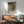 May include: Japanese woodblock print poster in wooden frame hanging on white wall above natural wood credenza. Print shows autumn landscape with orange and green trees along water, small figures on dock. Sage green upholstered chair with wooden arms positioned nearby, white ceramic table lamp, warm sunlight casting shadows across minimalist modern room setting.