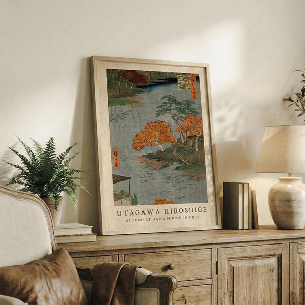 May include: Framed Japanese woodblock print reproduction featuring autumn landscape with orange and red foliage, river, and small figures, displayed on rustic wooden console alongside fern plant, books, and ceramic vase in neutral-toned living room setting