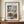 May include: Framed vintage Japanese woodblock print showing a snow-covered temple gate with traditional architecture, bare trees heavy with snow, and falling snowflakes against a dark blue sky. The artwork features muted colors of white, blue, brown and red. The print is displayed in a thin brown frame on a wooden credenza next to a small white ceramic vase and stacked books in a bright room with white walls and molding.
