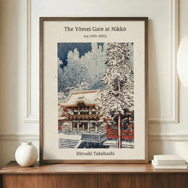 May include: Framed vintage Japanese woodblock print showing a snow-covered temple gate with traditional architecture, bare trees heavy with snow, and falling snowflakes against a dark blue sky. The artwork features muted colors of white, blue, brown and red. The print is displayed in a thin brown frame on a wooden credenza next to a small white ceramic vase and stacked books in a bright room with white walls and molding.