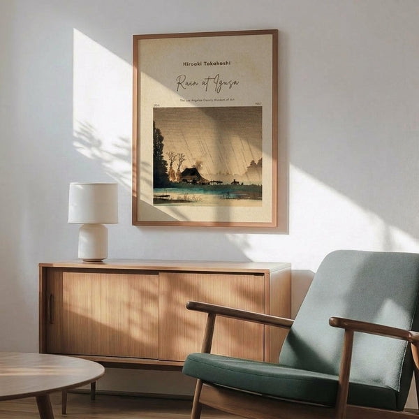May include: Framed vintage Japanese woodblock print showing a rainy landscape with traditional buildings, bare tree, and diagonal rain lines across sepia and teal tones. Displayed above a mid-century modern wooden credenza with white ceramic lamp, next to a sage green upholstered armchair with wooden frame in bright, naturally-lit room with white walls.