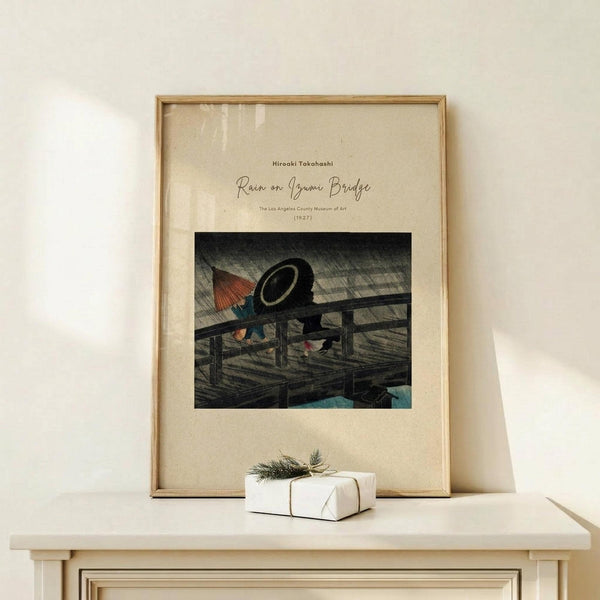 May include: Framed Japanese woodblock print poster displayed on light wood console table against cream wall. Artwork shows figures with umbrellas on wooden bridge in heavy rain, rendered in dark grays with touches of red and blue. Natural wood frame with cream matting. Small wrapped gift with twine and greenery sits beside frame. Soft natural lighting creates gentle shadows on minimalist setting.