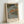 May include: Framed Japanese woodblock print reproduction showing autumn landscape with orange and green trees along a river, small figures on wooden walkway, traditional Asian text panels, displayed in light wooden frame leaning against white wall