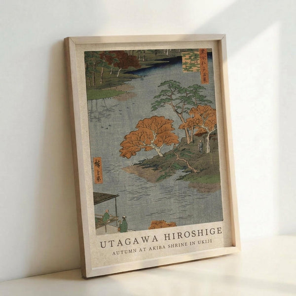 May include: Framed Japanese woodblock print reproduction showing autumn landscape with orange and green trees along a river, small figures on wooden walkway, traditional Asian text panels, displayed in light wooden frame leaning against white wall