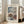 May include: Framed Japanese woodblock print showing a snow-covered temple gate with traditional architecture, bare winter trees, and falling snow against a dark blue sky. The vintage-style poster features muted colors with white snow contrasting against red temple elements. Light wooden frame leans against white wall next to beige upholstered chair in bright, minimalist living room with large windows and natural lighting.