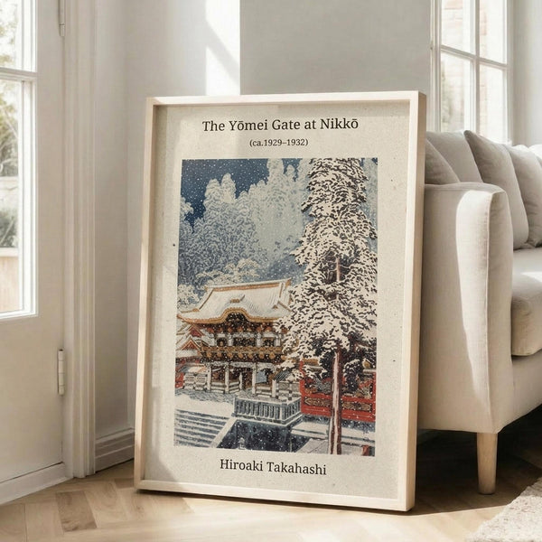 May include: Framed Japanese woodblock print showing a snow-covered temple gate with traditional architecture, bare winter trees, and falling snow against a dark blue sky. The vintage-style poster features muted colors with white snow contrasting against red temple elements. Light wooden frame leans against white wall next to beige upholstered chair in bright, minimalist living room with large windows and natural lighting.