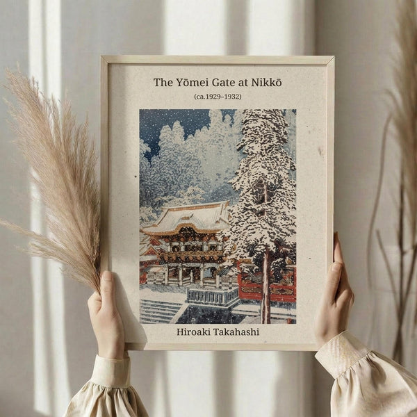 May include: Person holding framed vintage Japanese woodblock print showing snow-covered temple gate with traditional architecture, bare winter trees, and falling snow in muted blues and earth tones. Light wooden frame against neutral background with dried wheat stalks visible to the left side.