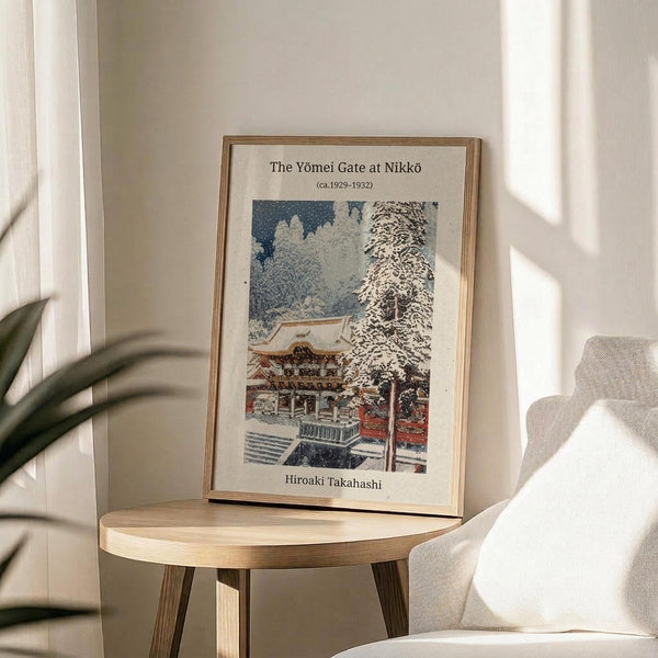 May include: Framed poster of a Japanese winter scene showing a snow-covered temple gate and buildings surrounded by bare trees under a deep blue sky. The vintage-style print features white text identifying it as "The Yōmei Gate at Nikkō" by Hiroaki Takahashi from 1929-1932. Display shows the artwork in a thin gold frame on a round wooden side table next to a cream-colored chair and plant, with natural sunlight streaming across white walls.
