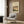 May include: Vintage Japanese woodblock print poster featuring a rainy landscape with traditional buildings, trees, and water under dramatic diagonal rain lines in muted earth tones. Shown framed in light wood on a textured wall above a cream upholstered armchair with geometric patterned pillow, beside a brass floor lamp and dark round side table with open book.