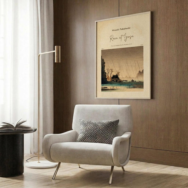 May include: Vintage Japanese woodblock print poster featuring a rainy landscape with traditional buildings, trees, and water under dramatic diagonal rain lines in muted earth tones. Shown framed in light wood on a textured wall above a cream upholstered armchair with geometric patterned pillow, beside a brass floor lamp and dark round side table with open book.