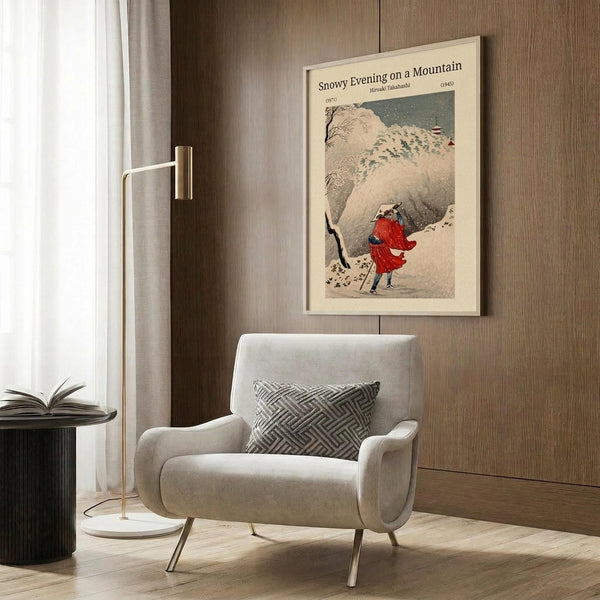 May include: Framed vintage Japanese woodblock print showing two figures in red robes walking through snowy mountain landscape with falling snow, displayed on wood-paneled wall above beige upholstered armchair with geometric patterned pillow, brass floor lamp and side table visible in modern neutral-toned room setting