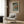 May include: Japanese woodblock print poster in white frame hanging on wood-paneled wall, showing autumn landscape with orange and green trees along a river. Cream upholstered armchair with geometric patterned pillow sits beneath artwork, accompanied by brass floor lamp and dark side table with open book. Natural lighting from nearby window illuminates the modern interior setting.