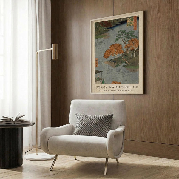 May include: Japanese woodblock print poster in white frame hanging on wood-paneled wall, showing autumn landscape with orange and green trees along a river. Cream upholstered armchair with geometric patterned pillow sits beneath artwork, accompanied by brass floor lamp and dark side table with open book. Natural lighting from nearby window illuminates the modern interior setting.
