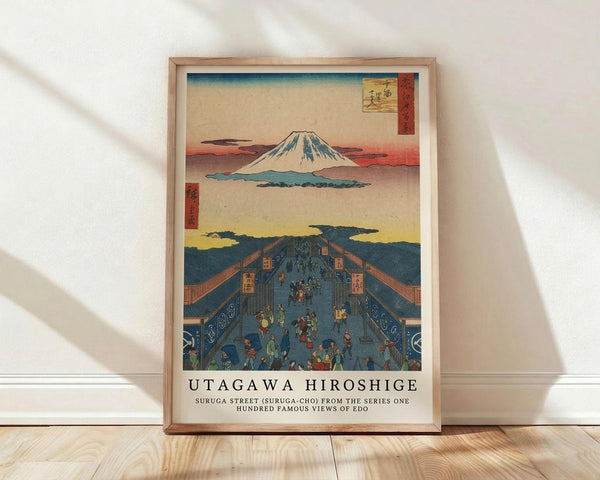 May include: Framed vintage Japanese woodblock print poster leaning against white textured wall on light wood floor. The artwork shows Mount Fuji in distance above a bustling blue street scene with small figures walking between wooden buildings. Warm sunset colors blend into cooler blues. Natural wood frame with white mat. Soft natural lighting creates diagonal shadows across the wall.