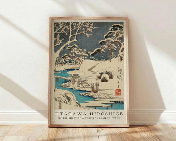 May include: Framed Japanese woodblock print featuring a snowy winter landscape with snow-covered pine trees, a stone lantern, and bright blue water stream. The artwork shows traditional ukiyo-e style with muted colors against a dark sky. Two figures in traditional dress walk along a snowy path. The print is displayed in a light wood frame leaning against a white wall on wooden flooring, with soft natural lighting creating diagonal shadows across the scene.