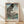 May include: Framed poster print of Japanese woodblock art featuring a large black and white scaled carp kite swimming above a landscape with Mount Fuji, traditional buildings, and a bridge. The artwork has soft blue-green skies and includes Japanese text characters. Natural wood frame displayed against white wall on light wooden flooring with window light casting diagonal shadows across the scene.