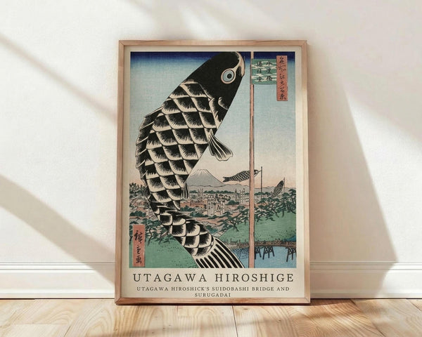 May include: Framed poster print of Japanese woodblock art featuring a large black and white scaled carp kite swimming above a landscape with Mount Fuji, traditional buildings, and a bridge. The artwork has soft blue-green skies and includes Japanese text characters. Natural wood frame displayed against white wall on light wooden flooring with window light casting diagonal shadows across the scene.