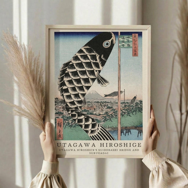 May include: Person in cream sweater holding framed Japanese woodblock print featuring large black carp kite with scaled pattern against blue sky, Mount Fuji and bridge landscape below. Light wood frame, dried pampas grass beside, soft window lighting creates warm interior styling scene.